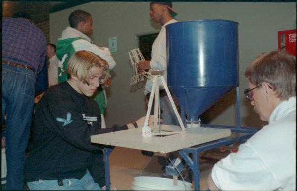 Two people kneeling at a table work on a science project. Two people in the background converse, with one holding a small constructed piece.