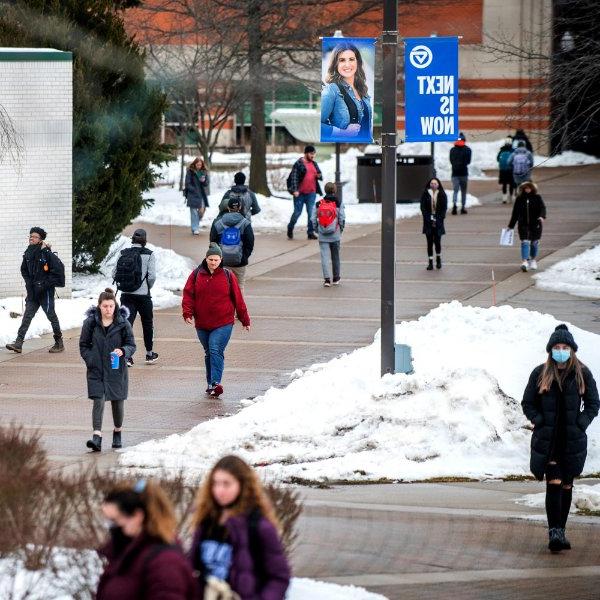 students walk on sidewalk, snow on grass areas; banner on lightpost reads Next is Now