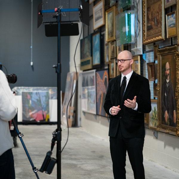 Nathan Kemler is interviewed for a video in a large room with lots of artwork behind him.